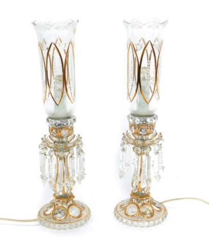 A pair of early 20thC Continental cut glass lustre table lamps, with white enamel floral decoration, gilt heightened, with brilliant and prismatic cut glass drops, and storm lantern shades, 53cm high.