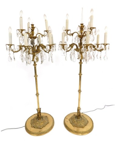 A pair of late 20thC brass two tier nine branch floor standing candelabra, with brilliant and tear shaped drops, raised on a foliate and scroll cast circular base, 164cm high.