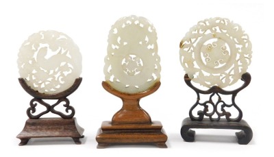 Three Chinese carved jade bi discs, one circular carved with a parrot and flowers, 5cm wide, another with flowers and leaves, 6cm wide, and an oblong disc decorated with a vase, framed by birds, 4.5cm wide, all on wooden stands.