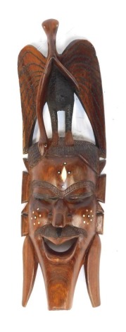 An African hardwood and resin inlaid wall mask, depicting a male face, surmounted by a bird holding snake in beak, 63cm high.