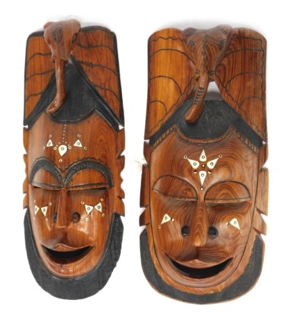 A pair of African hardwood and resin inlaid wall masks, each depicting a male face, surmounted by an elephant head, 60cm high.