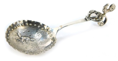 A Continental white metal caddy spoon, the bowl with a hammered edge and impressed beaded banding, decorated on an embossed scene depicting a figure on bench reaching towards child before building and fence, the handle decorated with a heart and scroll mo