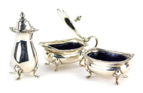 An Elizabeth II silver three piece cruet, comprising pepper pot, open salt, and lidded mustard pot, with blue glass liners, each with a gadrooned and shell capped border, raised on hoof supports, with salt spoon, weighable silver 4.84oz.