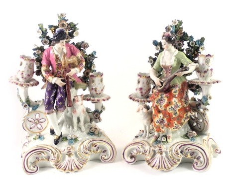 A pair of French Derby style porcelain candelabra, each modelled in the form of a male and female musician with lambs and dogs respectively, on a rococo scroll base, possibly Samson, 26cm high.