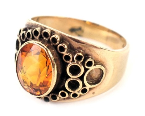 A 9ct gold Arts and Crafts style dress ring, with central oval cut citrine, in rub over setting, with outer applied moulded circle design, ring size P½, 7.3g all in.