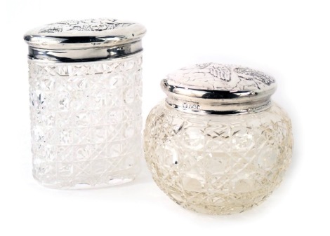 Two George V silver topped and pressed glass dressing table jars, one of oval design, with embossed cherubs to top, 9.5cm high, another of circular form, 7cm high, hallmark rubbed, Sheffield and Chester 1929 and later. (2)