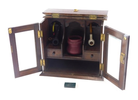 An early 20thC oak smoker's cabinet, with hinged lid and glazed doors, enclosing a small collection of pipes and a jar.