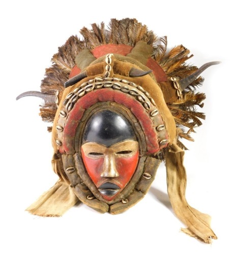 An African tribal Big Dan warrior mask, embellished with horns, shells, etc., with black, cream and red painted face and feather borders, Cote D'Ivoire, circa 1960, 50cm high.