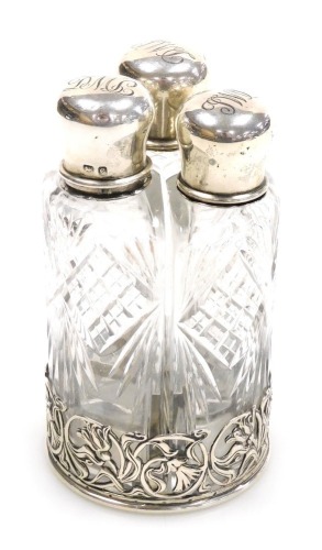 An Edward VII cut glass and silver set of three scent bottles, the lids monogram engraved, on a circular stand with pierced floral and foliate scroll decoration, William Hutton & Sons Limited, Birmingham 1902.