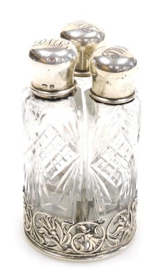 An Edward VII cut glass and silver set of three scent bottles, the lids monogram engraved, on a circular stand with pierced floral and foliate scroll decoration, William Hutton & Sons Limited, Birmingham 1902.