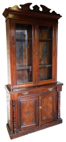 A Victorian mahogany cabinet bookcase, the break arch pediment over a pair of glazed doors enclosing three shelves, above a base with one long cushioned drawer over a pair of carved panel doors, raised on a plinth base united by a pair of turned pilasters