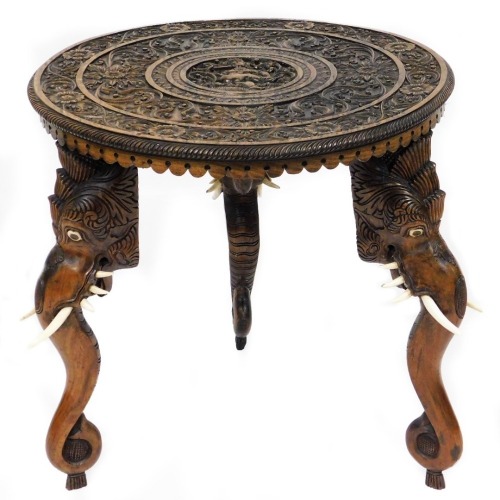 An early 20thC Anglo Indian hardwood occasional table, the circular top carved with the Hindu god Krishna, within inner and outer borders carved with repeating flowers and foliate scrolls, raised on three elephant's head legs, each set with two ivory tusk