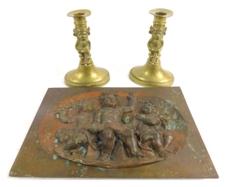 A copper plaque, relief mould decorated with three seated cherubs, registration mark verso, 26cm x 17cm, together with two brass candlesticks decorated with a figure of the Lincoln Imp, each 15cm high.