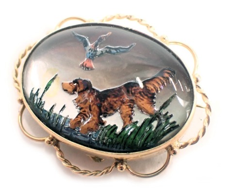 An oval animal brooch, with mother of pearl back, raised painted decoration of terrier and ducks in reeds, with looped outer border, 3.5g diameter, yellow metal stamped 9ct, with single pin back and safety chain, 11.8g all in.