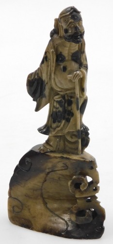 A late 19thC Chinese soapstone figure of an Immortal, carved standing with an exposed rib cage and distended belly, holding a stick, raised on a carved base, 18cm high.