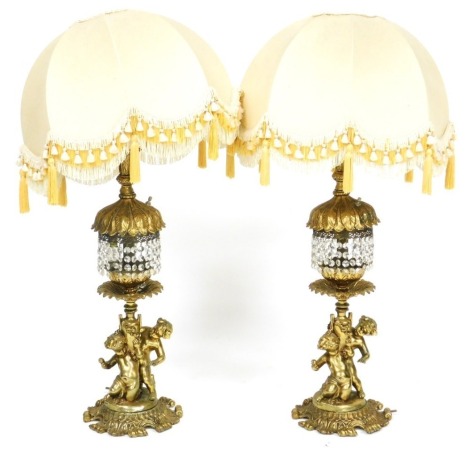 A pair of 20thC Continental brass table lamps, modelled with putti, on a scrolling leaf base, with brilliant cut glass drops and shades, 91cm high. Buyer Note: WARNING! This lot contains untested or unsafe electrical items. It is supplied for scrap or re