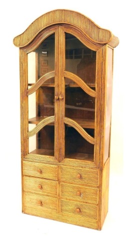 A cane reeded display cabinet, with an arched top above two panelled doors and six short drawers, 205cm high, 87cm wide, 40cm deep.