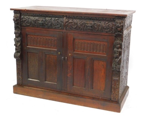 A Victorian carved oak bookcase base cupboard, fitted with two drawers and enclosed by two panelled doors, carved with lions heads and cherubs in relief, 98cm high, 125cm wide.