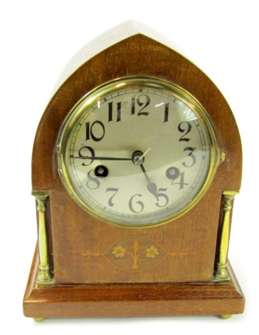 An early 20thC mahogany cased mantel clock, of arched form, the circular silvered dial bearing Arabic numerals, eight day movement, the case with an inlaid panel depicting a flowering tree, flanked by brass columns, on a stepped plinth base, raised on fou