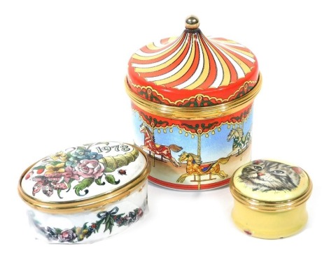 A Halcyon Days enamel musical box, The Carousel Waltz, together with a rouge pot, decorated to the lid with a cat, and an oval box and cover for 1978, decorated with a cornucopia of flowers. (3, AF)