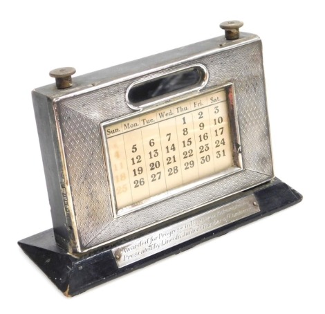 An Elizabeth II silver faced desk calendar, engine turned, with paper central label and twin dials, mounted on a black painted base bearing presentation plaque Awarded For Progress In Business Administration Presented By Lincoln Junior Chamber of Commerce