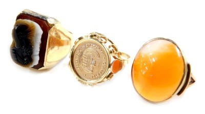 Three dress rings, comprising intaglio headed gentleman's signet ring, yellow metal, unmarked, size T 1/2, an oval orange agate dress ring, V splayed shoulders, yellow metal, unmarked, size M, and a coin dress ring, marked with Egyptian figures, pierced m