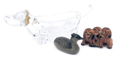 A Vannes le Chatel glass trinket dish, modelled as a dachshund, partial paper label, 26cm wide, together with a Poole pottery figure of grebe, and a Beswick pottery figure group of three spaniels, printed marks, 9cm wide.