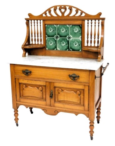 A Victorian marble topped satin walnut washstand, with a carved crest rail, above a six green floral tiled splash back flanked by turned spindles and two quarter shaped shelves, over a long drawer above a pair of cupboard doors, raised on turned legs, on 