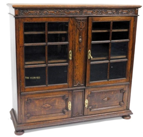 An Edwardian oak display cabinet, with carved frieze to top, above two glass panelled doors, over two cupboards below, on bun feet, 122cm high, 128cm wide, 41cm deep.