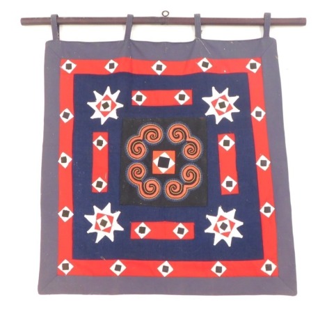 Tribal Art. An embroidered panel, on a navy ground with red star border, mahogany pole, 79cm high, 81cm wide.