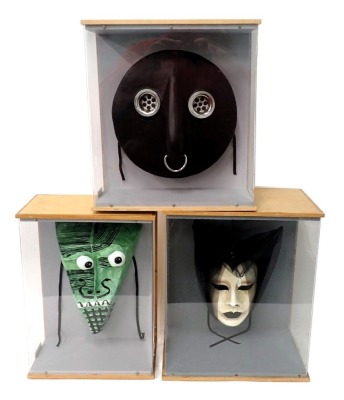 A collection of three masks, a Venetian mask with veil, a fetish plug mask, and an abstract papier mache face mask decorated in green, each in a glazed perspex case.