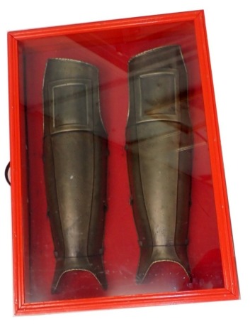 Two replica armour leg guards, in red painted and perspex case, 75cm wide.