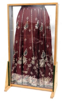 A red silk skirt, embroidered with design of peacocks, in metallic thread, mounted into a floor standing perspex and wooden display case, 108cm high, 64cm wide.