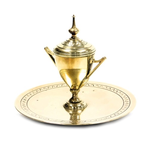 A Victorian brass inkwell, of trophy form, with a ceramic inset, raised on a circular base, 16.5cm wide.