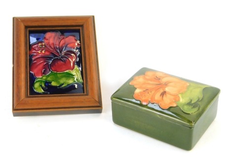 A Moorcroft pottery trinket dish and cover, of rectangular form, decorated in the Green Hibiscus pattern, impressed marks, 9.5cm wide, together with a pot lid or plaque, later framed, 12cm x 9.5cm. (2)