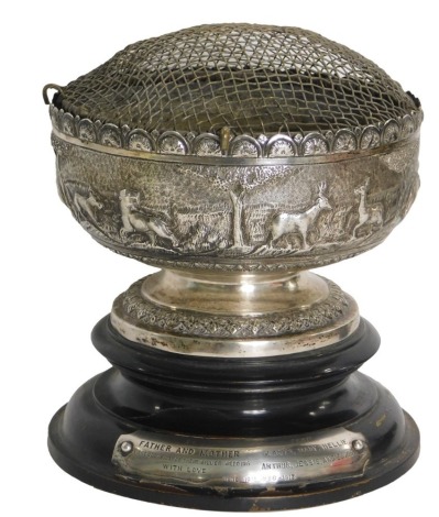 An Indonesian white metal bowl, with hammered safari scene body, on a stepped and ribbed foot, with later mesh insert, on an ebonised base with silver plated presentation plaque dated 1913, the bowl 8.14oz, 18cm high on base.