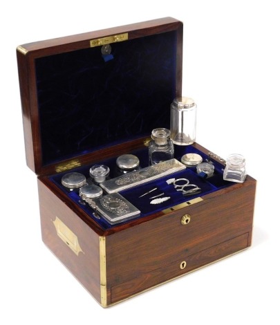 A Victorian rosewood and brass bound travelling box, the hinged lid opening to reveal a purple velvet fitted interior with a lift out tray containing cut glass and plate lidded bottles, boxes and jars, three manicure implements, with five recesses behind 