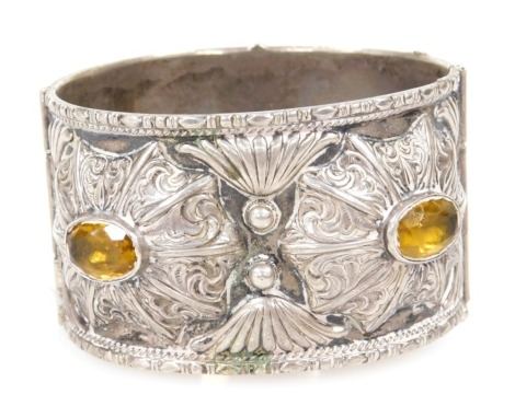 A Continental bangle, white metal, embossed in a floral and foliate design, set with four yellow stones, possibly citrines, with a pin clasp, 76.9g all in.