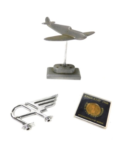 A vintage Austin chrome bonnet badge, the Flying A, together with a World Cup Championship Willie commemorative medal, 1966 and pewter desk stand modelled as a spitfire, 11.5cm high. (3)