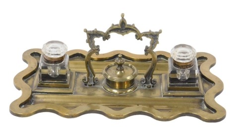 A Victorian brass desk stand, of wavy edge form, with scrolling carrying handle, and a pair of cut glass inkwells with hinged stoppers, 31cm wide.