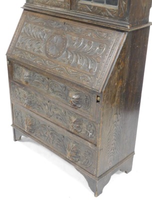 A 1920's oak bureau bookcase, with lion's mask handles, and profusely floral and foliate carved, the plain pediment over a pair of glazed doors, internal shelves lacking, above a fall flap opening to reveal a single drawer and six recesses, over three gra - 2