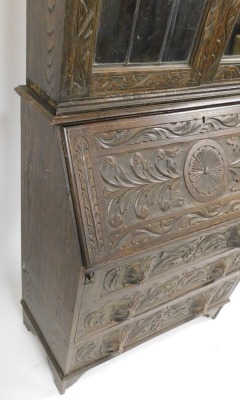 A 1920's oak bureau bookcase, with lion's mask handles, and profusely floral and foliate carved, the plain pediment over a pair of glazed doors, internal shelves lacking, above a fall flap opening to reveal a single drawer and six recesses, over three gra - 3