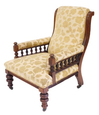 A Victorian mahogany armchair with galleried arms, and overstuffed back, seat and arm rests, in gold floral and foliate fabric, raised on turned legs, on castors, 67cm wide.