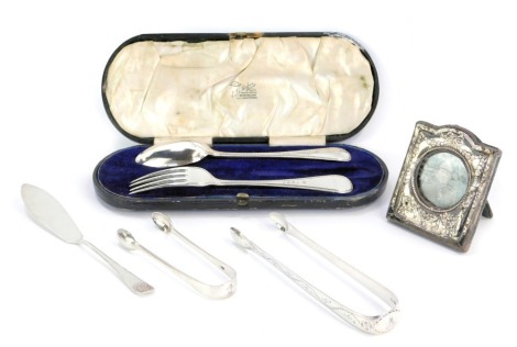 Silver cutlery and effects, comprising a child's silver spoon and fork set, with beaded border, bearing the initials JD, a silver framed photograph frame, pair of Elizabeth II silver engine engraved pattern sugar tongs, a miniature cake slice, and a plain