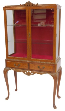 A 20thC burr walnut display cabinet in Queen Anne style, the top with a scroll carved frieze above two glass doors enclosing glass shelves, with glass side panels, the base with two drawers above a scroll carved apron, on carved cabriole legs and pad feet