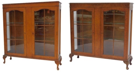 A pair of 20thC burr walnut display cabinets in Queen Anne style, each top with a carved edge above two glazed panelled doors enclosing two glass shelves, with glass side panels, with a moulded base raised on carved legs with pad feet, 116cm high, 109cm w