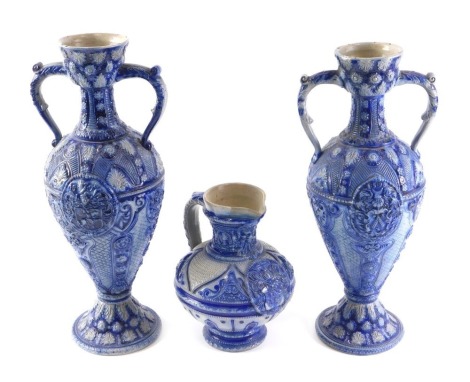 A pair of German blue glazed stoneware two handled vases, each decorated in relief with a crest, roundels, etc., 38cm high, and a similar jug, 20cm high.