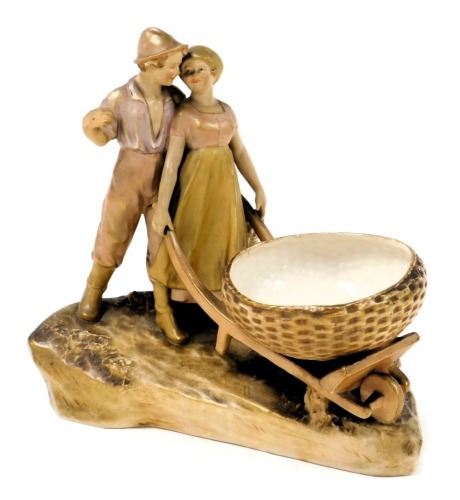 A Royal Dux blush porcelain figural bowl, modelled as a man alongside a lady pushing a wheelbarrow, raised on a naturalistic base, number 235, impressed and raised triangle marks, 29cm wide.