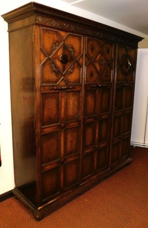 A late Victorian oak compactum wardrobe, by James Shoolbred of London, the outswept pediment with dentil moulding, above a floral and foliate carved frieze, above three carved panelled doors, opening to reveal a shelf over a hanging rail and hooks, flanke