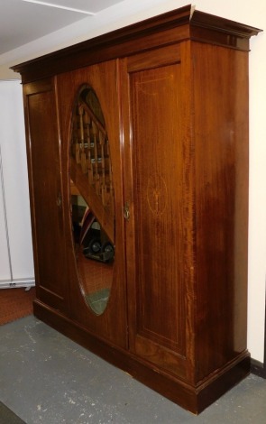 An Edwardian mahogany and inlaid triple wardrobe, the outswept pediment over a central oval bevelled glass mirror plate, flanked by two doors with inlaid cornucopia and bow tied harebell decoration, opening to reveal a hanging rail and hooks, above one lo